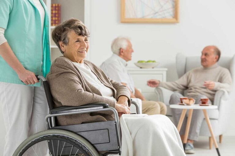 An older adult embraces their visitor in a warmly lit assisted living common room.